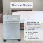 A wheeled bedroom air cleaner shown in a bedroom setting with a bed in the background. The unit has a light gray top section with a dial and a darker gray lower mesh body.
